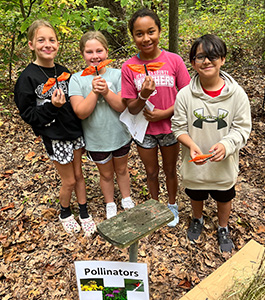 students outside in front of a sign about pollinators. They are holding orange butterflies they have made